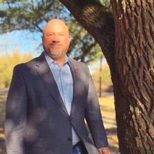 Man in grey blazer and blue jeans standing next to tree.